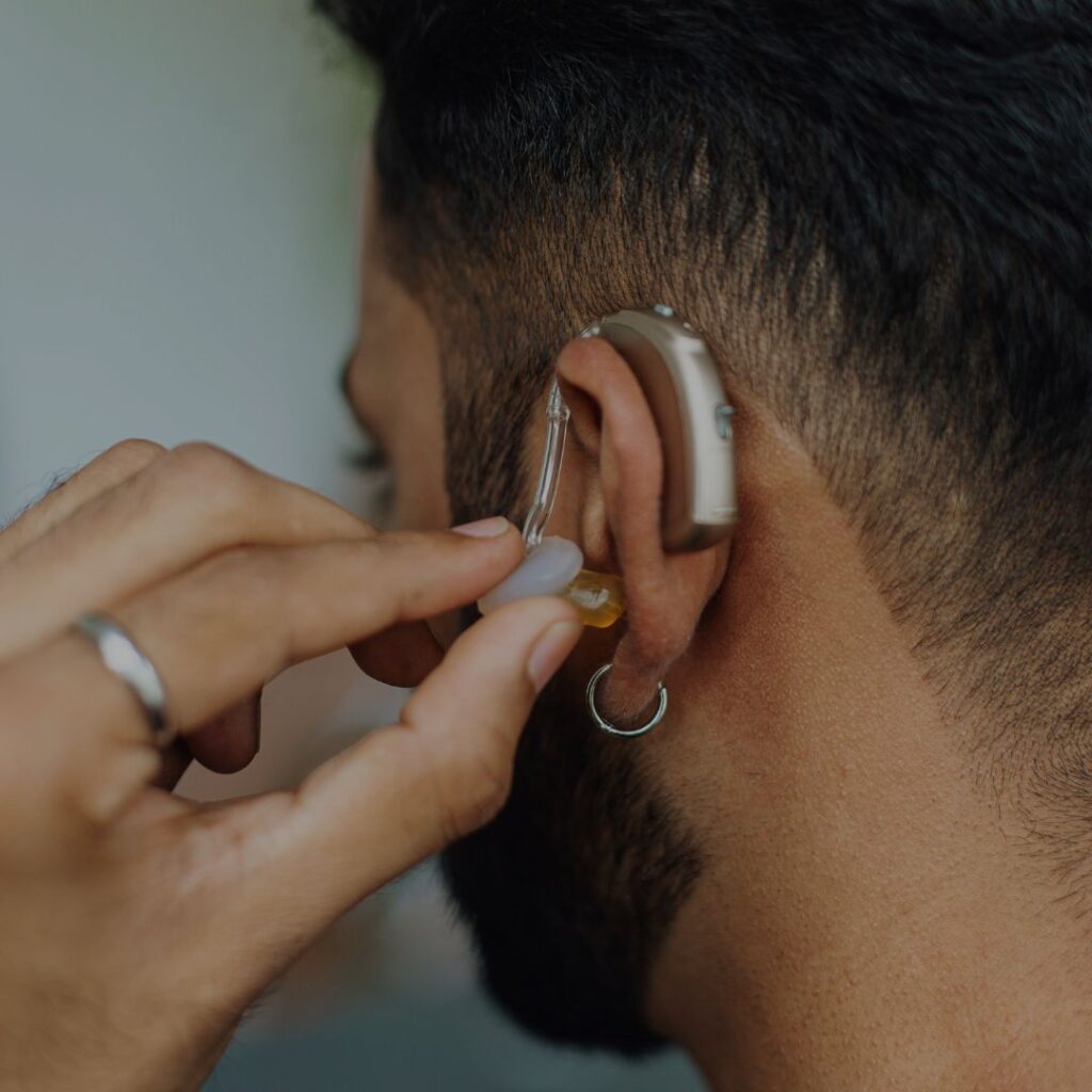 Bearded man facing away placing a hearing aid in his left ear which has a piercing. 